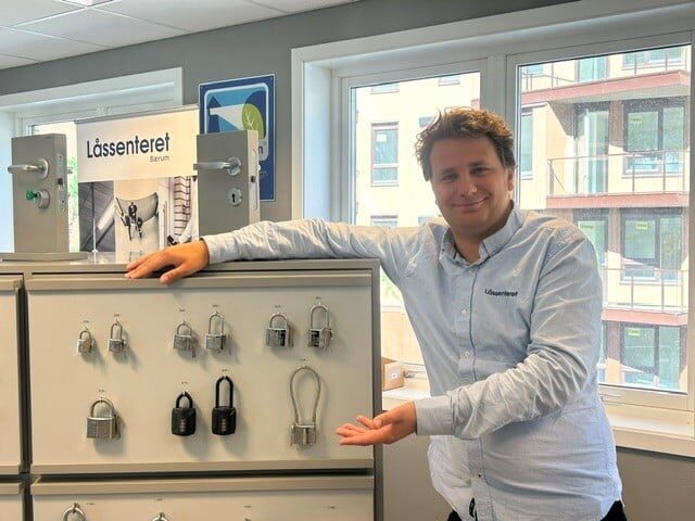 Hengelåser Bærum En mann står innendørs, ved siden av en utstilling av ulike hengelåser montert på metallskap, med vinduer og boligblokker i bakgrunnen.