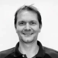 Morten-300x300 En mann med kort hår smiler mot kameraet. Han er iført en mørk skjorte med krage, og er plassert mot en enkel, lys bakgrunn.