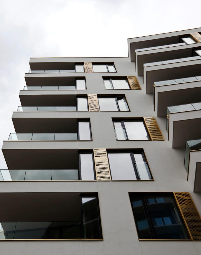 Moderne boligblokk med flere balkonger, glassrekkverk og geometriske arkitektoniske detaljer, sett fra en lav vinkel mot en overskyet himmel.