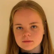 En ung person med langt, blondt hår og nøytralt ansiktsuttrykk står vendt mot kameraet mot en ensfarget, lys bakgrunn.