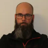 En mann med skallet hode, briller og langt helskjegg har på seg en mørk genser med glidelås og ser direkte inn i kameraet mot en ensfarget bakgrunn.