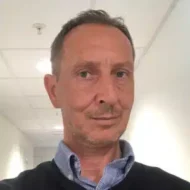 Pal-Ringvold-300x300 En mann med kort hår og trimmet skjegg, iført blå skjorte med krage og mørk genser, står i en opplyst gang og ser inn i kameraet.