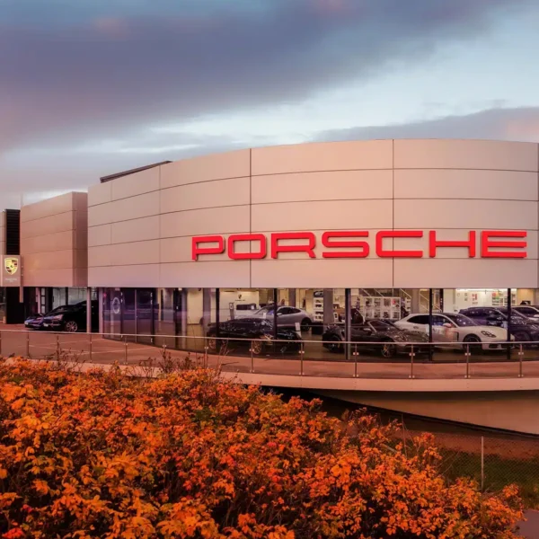 Moderne Porsche-forhandlerbygning med store glassvinduer, en fremtredende rød Porsche-logo og flere utstilte biler; høstløv i forgrunnen.