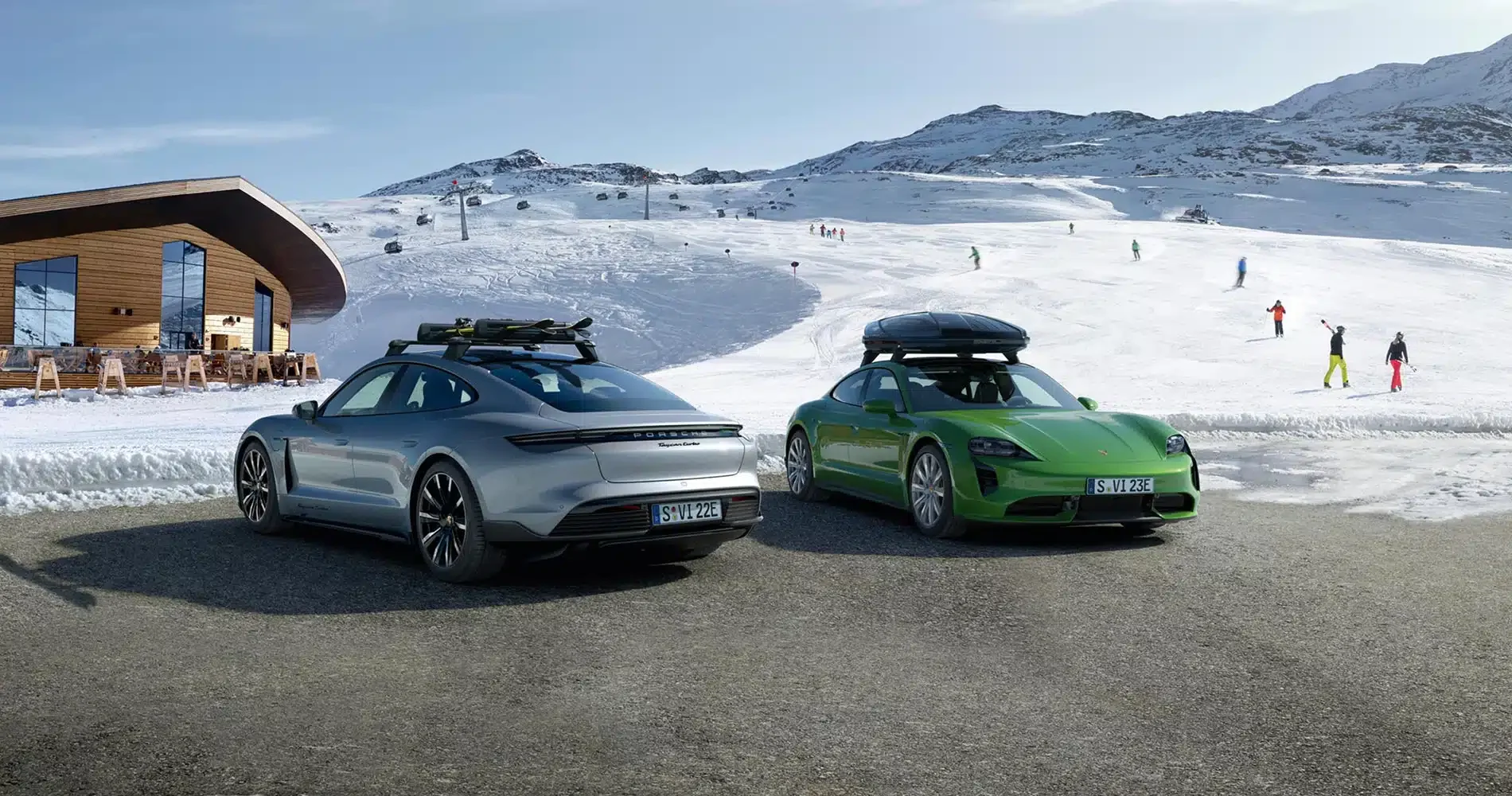 To elbiler med takstativ står parkert i nærheten av en moderne bygning, med snødekte fjell og folk på ski i bakgrunnen.