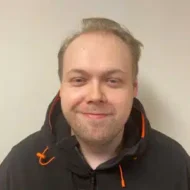 Rayner-300x300 En mann med lys hud og kort, lysebrunt hår er iført en svart jakke med oransje aksenter. Han står mot en beige vegg og smiler lett.