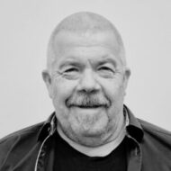 En middelaldrende mann med kort, grått hår og skjegg, iført mørk skjorte over en svart t-skjorte, smiler til kameraet mot en vanlig, lys bakgrunn.