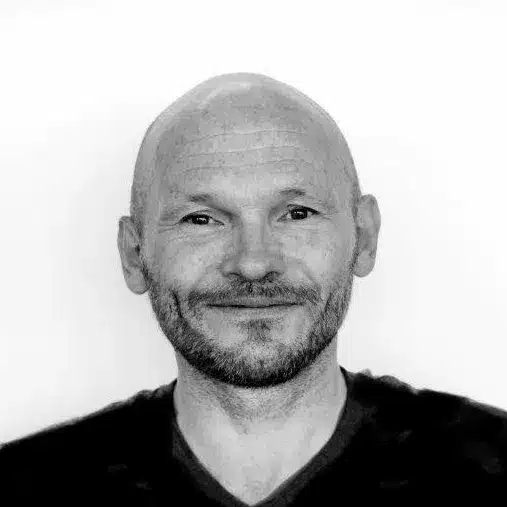 Ronny En skallet mann med skjegg og bart smiler lett til kameraet i et svart-hvitt portrett mot en ensfarget bakgrunn.