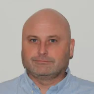 En middelaldrende mann med barbert hode, lys hud og lyse skjeggstubber, iført en lyseblå skjorte med krage, står vendt mot kameraet mot en ensfarget hvit bakgrunn.