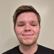 Sander Fladvad En ung mann med kort brunt hår og lett ansiktshår ser inn i kameraet og smiler litt, mens han står foran en ensfarget, lys bakgrunn.