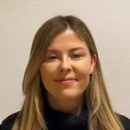 Sandra-300x300 En kvinne med rett, lysebrunt hår og et nøytralt uttrykk står foran en beige vegg, iført en svart topp.