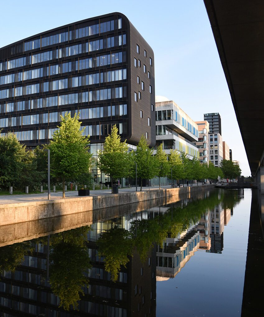 Moderne kontorbygninger og trær står langs en kanal, og speilingene av dem er synlige i det rolige vannet under en klar himmel.