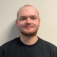 Simen-Ludvigsen-e1712135637209-300x300 En person med kort hår og trimmet skjegg, iført svart genser, står foran en ensfarget, lys vegg og smiler lett til kameraet.