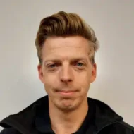 En mann med lys hud og blondt hår som er stylet opp, står vendt mot kameraet mot en ensfarget, lys bakgrunn.