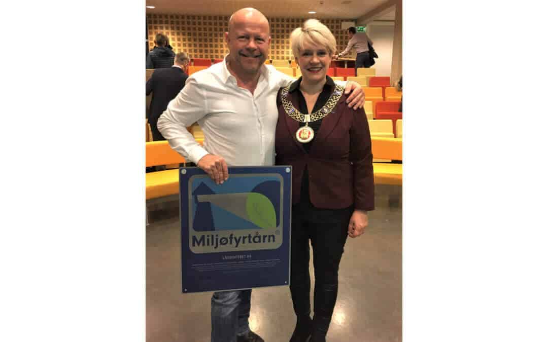 To personer står i et auditorium, den ene holder en Miljøfyrtårn-plakett. Den andre personen er kledd i et formelt antrekk med en dekorativ medaljong.