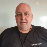 En middelaldrende mann med kort, grått hår og skjegg, iført en svart skjorte med påskriften "Låssenteret", står mot en enkel, hvit bakgrunn.