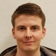 En ung mann med kort, brunt hår og et lite smil står vendt mot kameraet mot en lys, strukturert vegg.