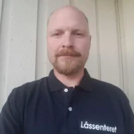 Stian-B-Drammen-e1599463598379 En mann med skjegg og bart, iført svart skjorte med teksten "Låssenteret", står foran en lys trevegg.