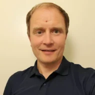 En mann med kort, lysebrunt hår og marineblå skjorte med krage smiler til kameraet mot en ensfarget, lys bakgrunn.