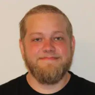 En mann med lys hud, kort blondt skjegg og kort hår smiler lett til kameraet mot en vanlig lys bakgrunn.
