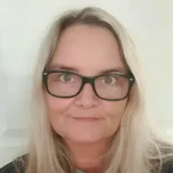 En kvinne med langt, blondt hår og svarte briller ser inn i kameraet med et nøytralt uttrykk. Hun står foran en ensfarget, lys bakgrunn.