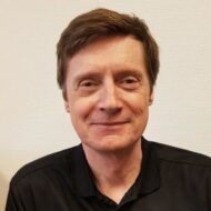 En middelaldrende mann med kort, brunt hår og svart skjorte med krage smiler til kameraet mot en ensfarget, lys vegg.