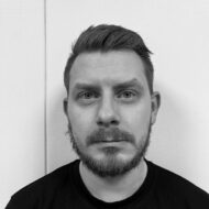 En mann med kort hår og skjegg ser direkte inn i kameraet mot en vanlig, strukturert vegg; bildet er i svart-hvitt.