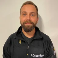 Thomas-Lunde-rotated-e1599548765778 En mann med skjegg og bart står mot en ensfarget, lys vegg, iført en svart jakke med påskriften "Låssenteret".