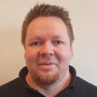 ThomasS En mann med kort, brunt hår med pigger og rødblondt fippskjegg er iført svart skjorte og står vendt mot kameraet mot en lys vegg med tekstur.