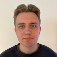 En ung person med kort, lysebrunt hår og blå øyne ser rett inn i kameraet foran en ensfarget hvit bakgrunn.