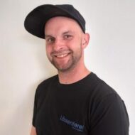 En mann i svart t-skjorte og svart caps smiler til kameraet mot en hvit bakgrunn.