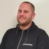 En mann med kort hår og skjegg, iført en mørk hettegenser med påskriften "Låssenteret", står mot en ensfarget, lys bakgrunn.