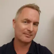 En mann med kort, lyst hår og et lite smil har på seg en mørk skjorte og står vendt mot kameraet mot en enkel, lys bakgrunn.