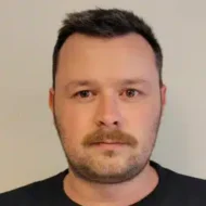 En mann med kort, mørkt hår, bart og skjeggstubber ser direkte inn i kameraet mot en vanlig, lys bakgrunn.