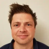 En mann med kort, brunt hår med pigger og et lite smil står vendt mot kameraet mot en ensfarget, lys bakgrunn.