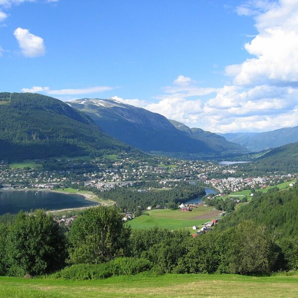 En by som ligger i en dal mellom skogkledde fjell og en innsjø under en blå himmel.