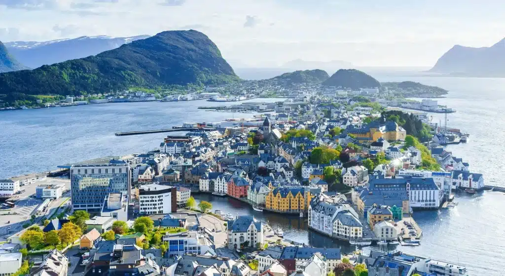 aalesund Flyfoto av en kystby med fargerike bygninger, omgitt av vann og fjell under en delvis overskyet himmel.