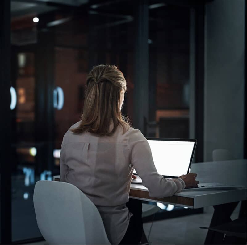 En person sitter alene ved et skrivebord og jobber på en datamaskin på et dunkelt kontor om kvelden.