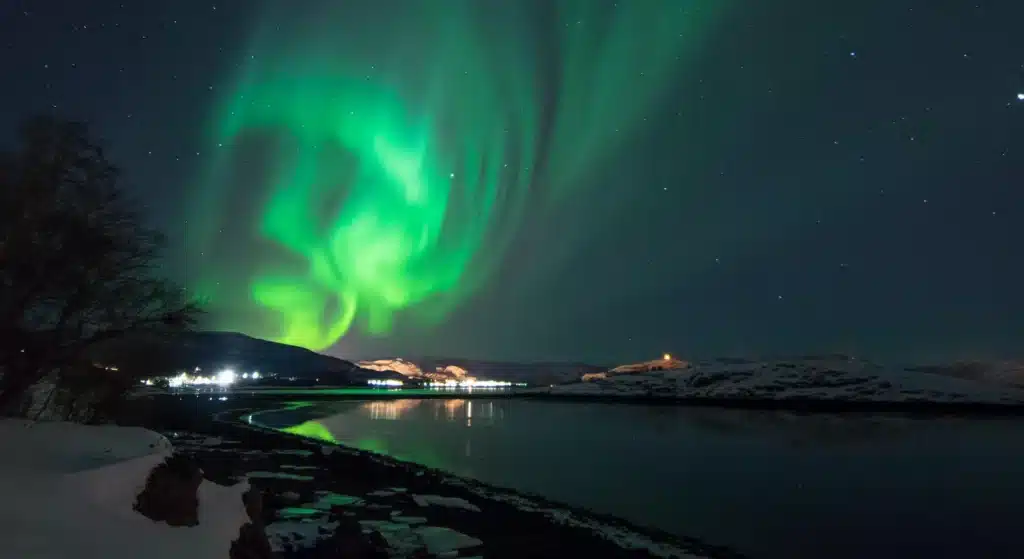 Grønne nordlys hvirvler rundt på nattehimmelen over et snødekt landskap, og reflekterer seg i et stille vann med bygninger i det fjerne.