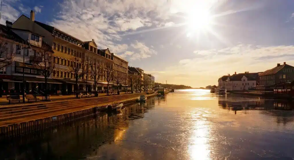En kanal renner gjennom en by med mange bygninger under en strålende sol og delvis overskyet himmel, med speilinger i vannet og tomme uteserveringer.