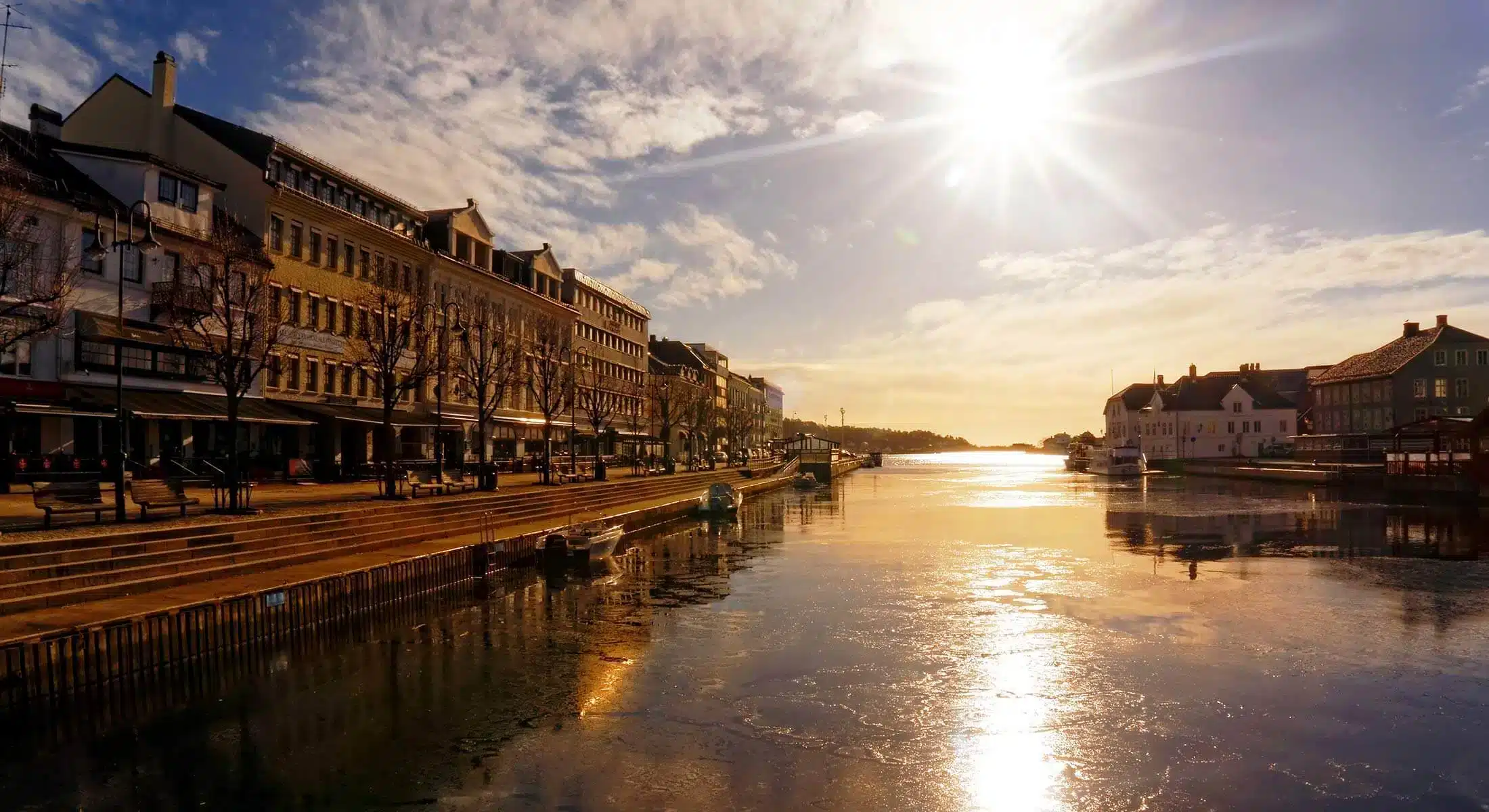 En kanal renner gjennom en by med mange bygninger under en strålende sol og delvis overskyet himmel, med speilinger i vannet og tomme uteserveringer.