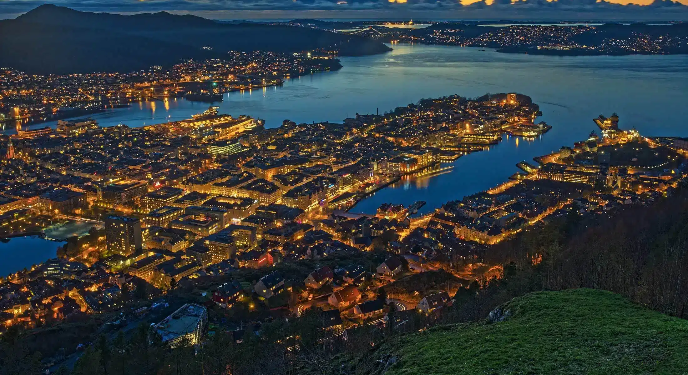 Flyfoto av en kystby i skumringen med opplyste gater og bygninger ved et vann, omgitt av åser.