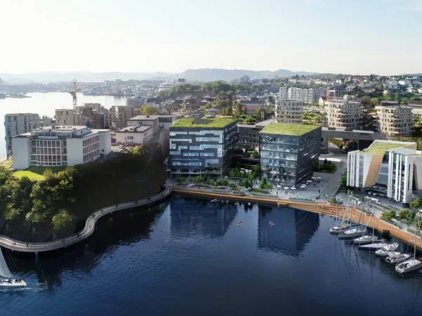 Flyfoto av et moderne bylandskap ved vannet med bygninger med grønne tak, en marina med båter ved kai og en seilbåt på rolig vann.