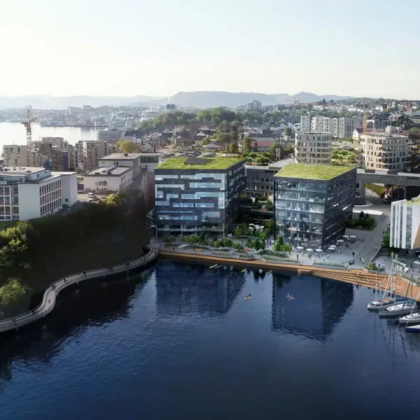 Flyfoto av et moderne bylandskap ved vannet med bygninger med grønne tak, en marina med båter ved kai og en seilbåt på rolig vann.