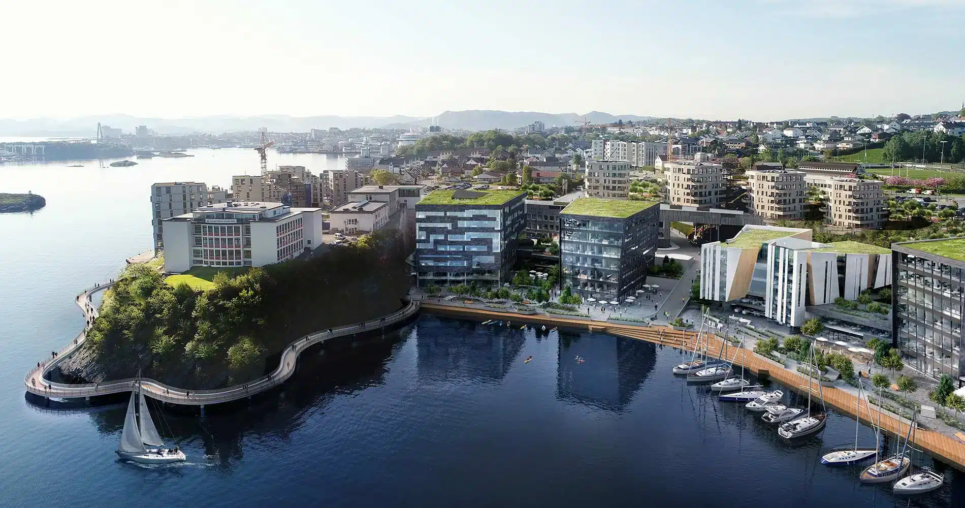 Flyfoto av et moderne bylandskap ved vannet med bygninger med grønne tak, en marina med båter ved kai og en seilbåt på rolig vann.