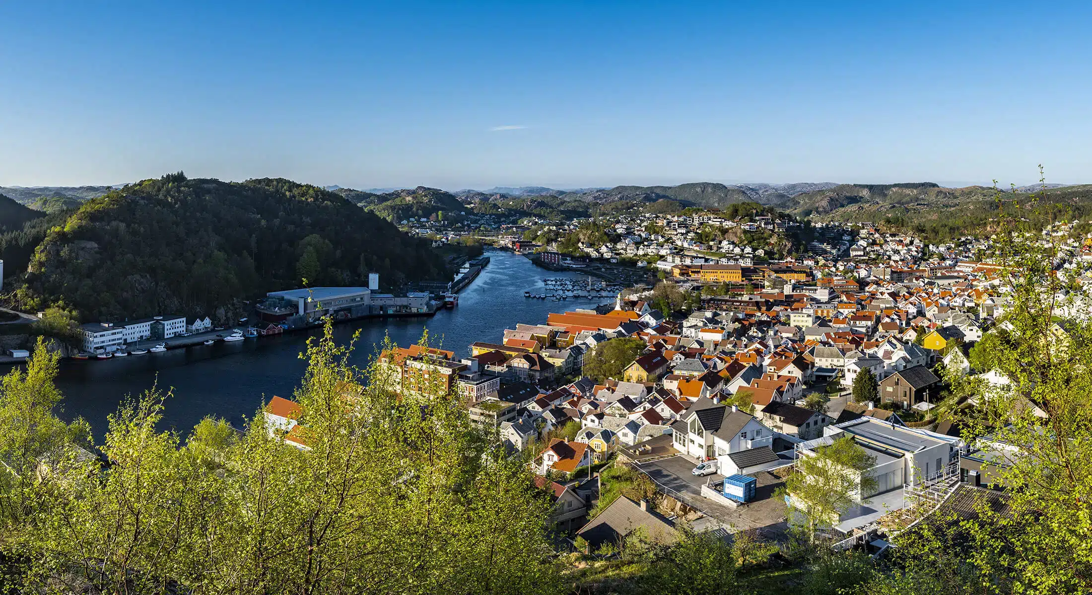 Panoramautsikt over en elveby med røde hustak, grønne åser og en elv som slynger seg gjennom landskapet under en klarblå himmel.