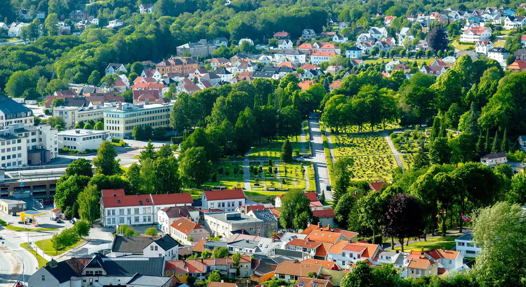 Flyfoto av en by med røde hus, grønne trær og en kirkegård, omgitt av tett skog.