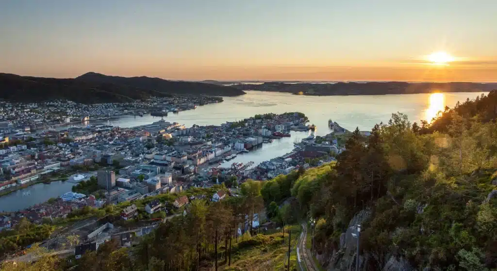 Et bylandskap med bygninger ved vannkanten og grønne åser i solnedgang, sett fra et høyt utsiktspunkt over en bukt.