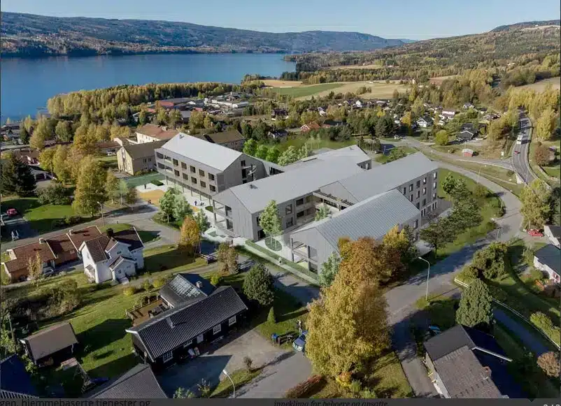 Flyfoto av et moderne bygningskompleks under oppføring i et forstadsområde nær en innsjø, omgitt av hus, trær og veier.