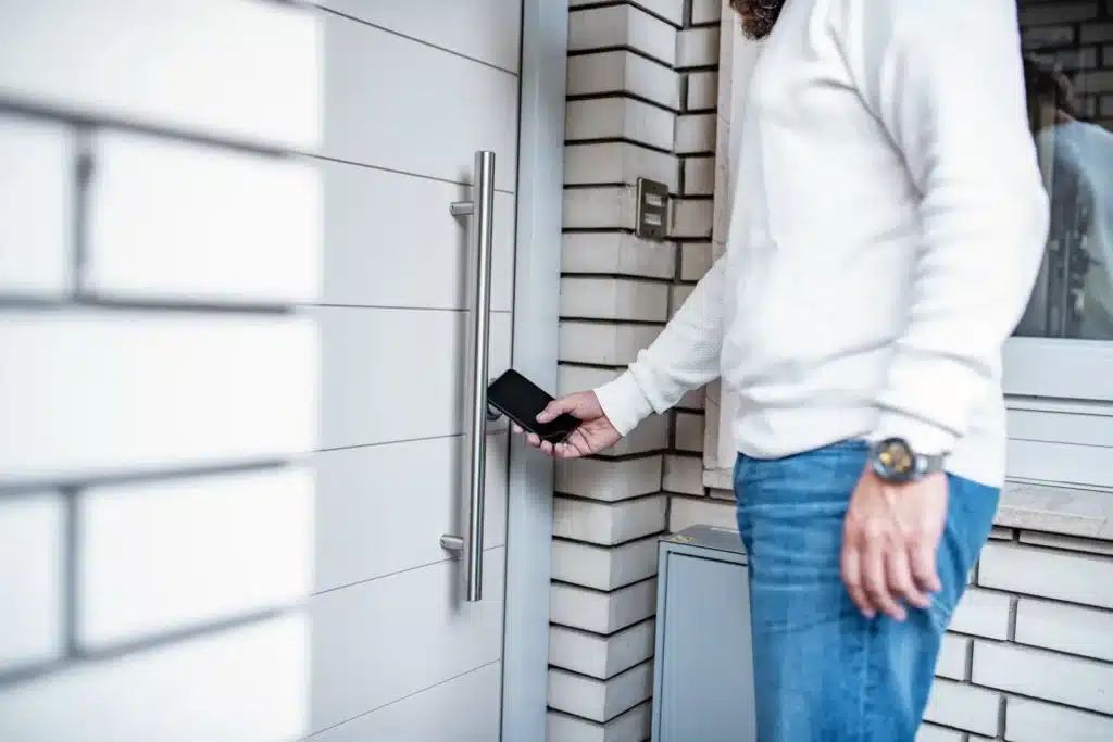 En person holder en smarttelefon i nærheten av en moderne inngangsdør, muligens for å låse opp døren via en elektronisk lås og smartteknologi.