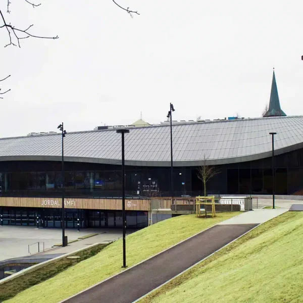 Bak en gressbakke og asfalterte gangveier ligger en moderne bygning med et bølgende, grått tak og store glassvinduer en overskyet dag.